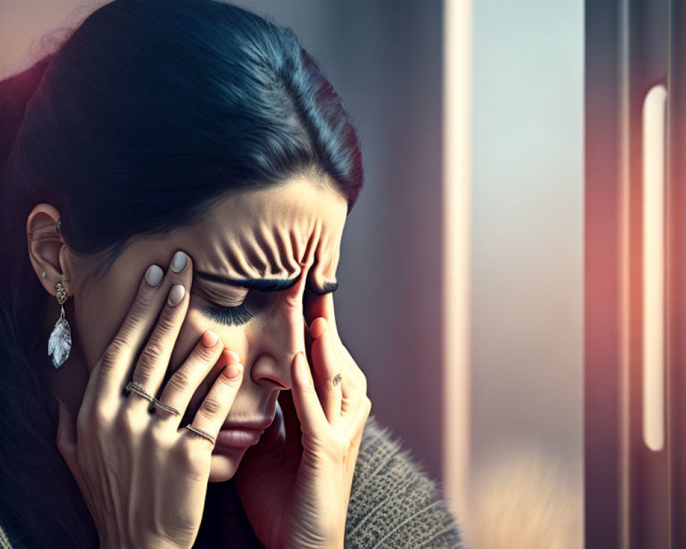 Distressed woman with long hair in anguish holding head, blurred window background