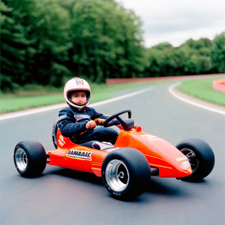 Child in racing suit and helmet drives orange go-kart on outdoor track