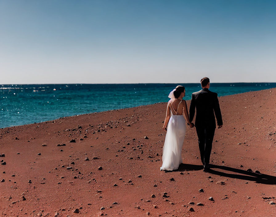 Bride and groom in white dress and black suit walking on pebbly beach by blue sea