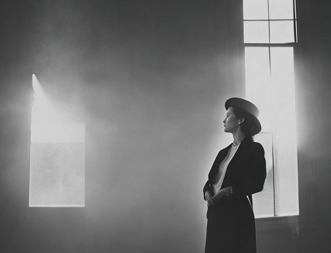 Woman in hat and coat gazes at light in hazy room