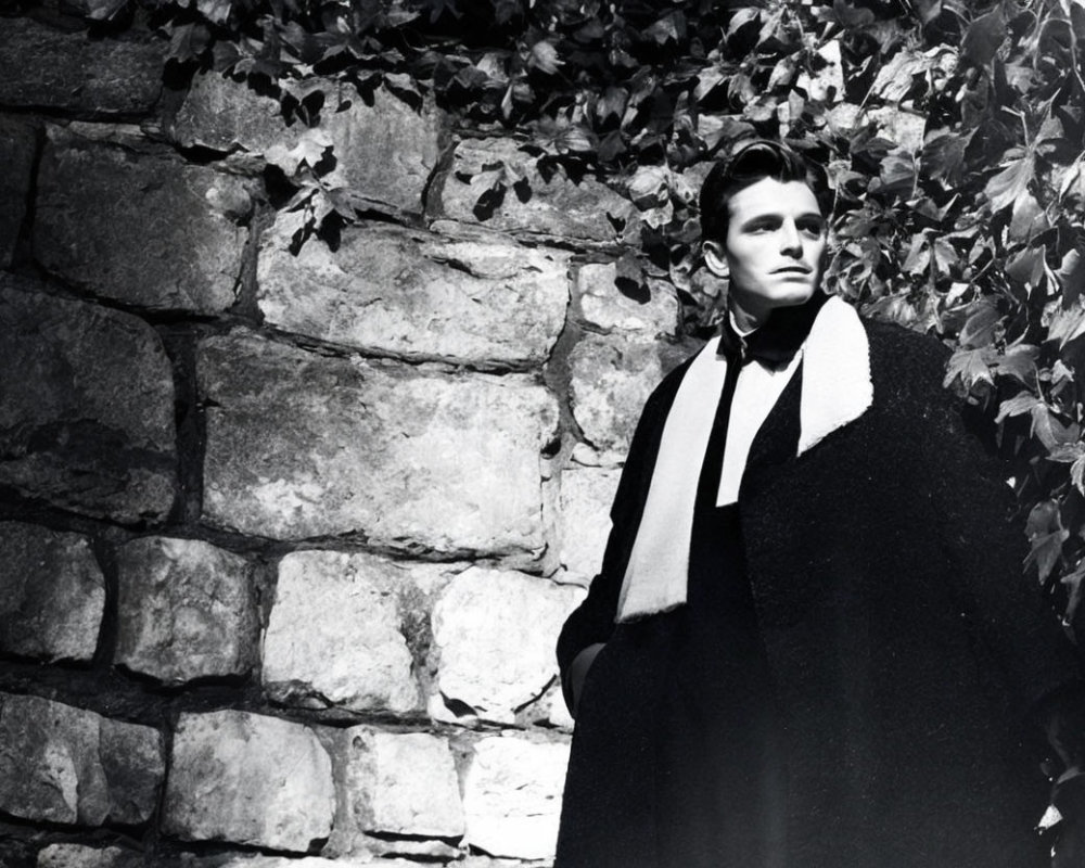 Vintage-clad man in cape against vine-covered wall in black and white.