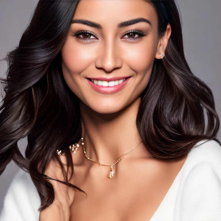 Smiling woman with long dark hair in white top and necklace on grey background