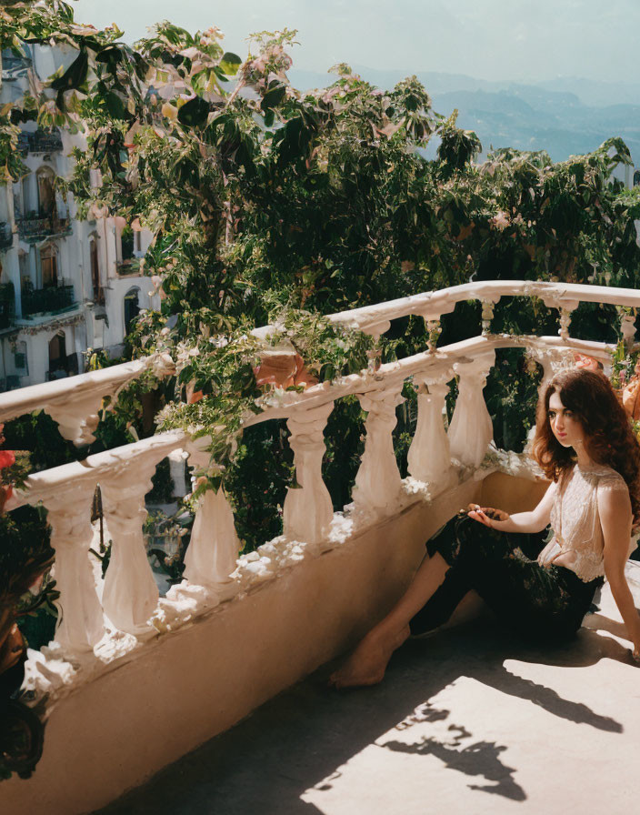 Woman sitting on sunny balcony with ornate balustrades, overlooking lush cityscape.