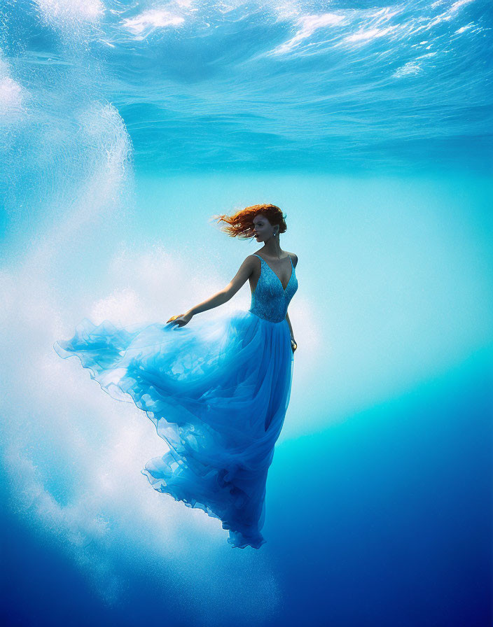 Woman in flowing blue dress underwater with red hair backlit by sunlight