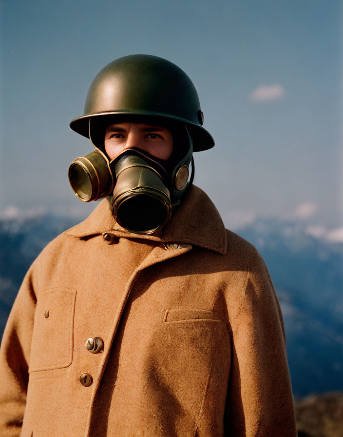 Military person in helmet and gas mask against mountain backdrop in brown overcoat