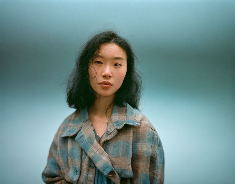 Dark-haired young woman in plaid shirt against soft blue backdrop