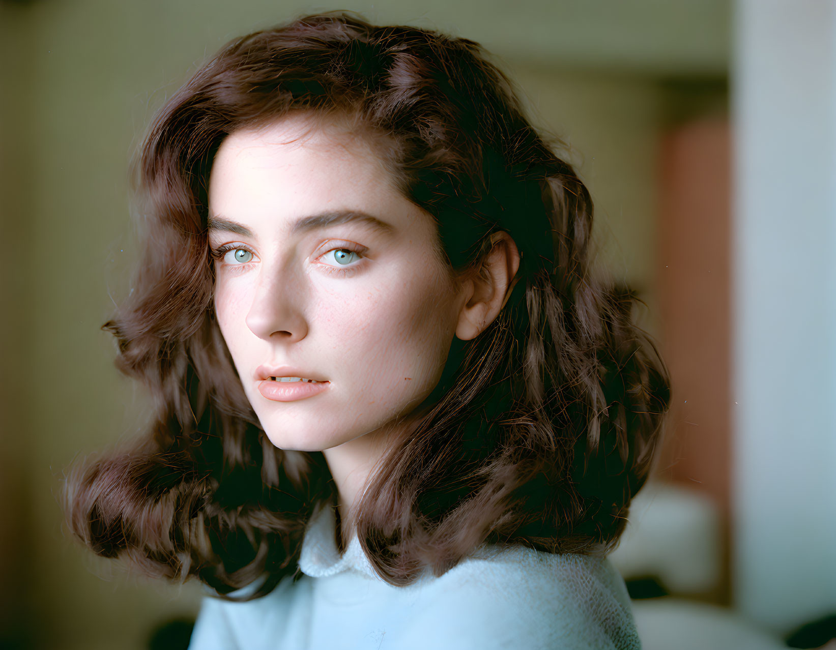 Young woman with wavy brown hair and blue eyes in light blue sweater gazes at camera