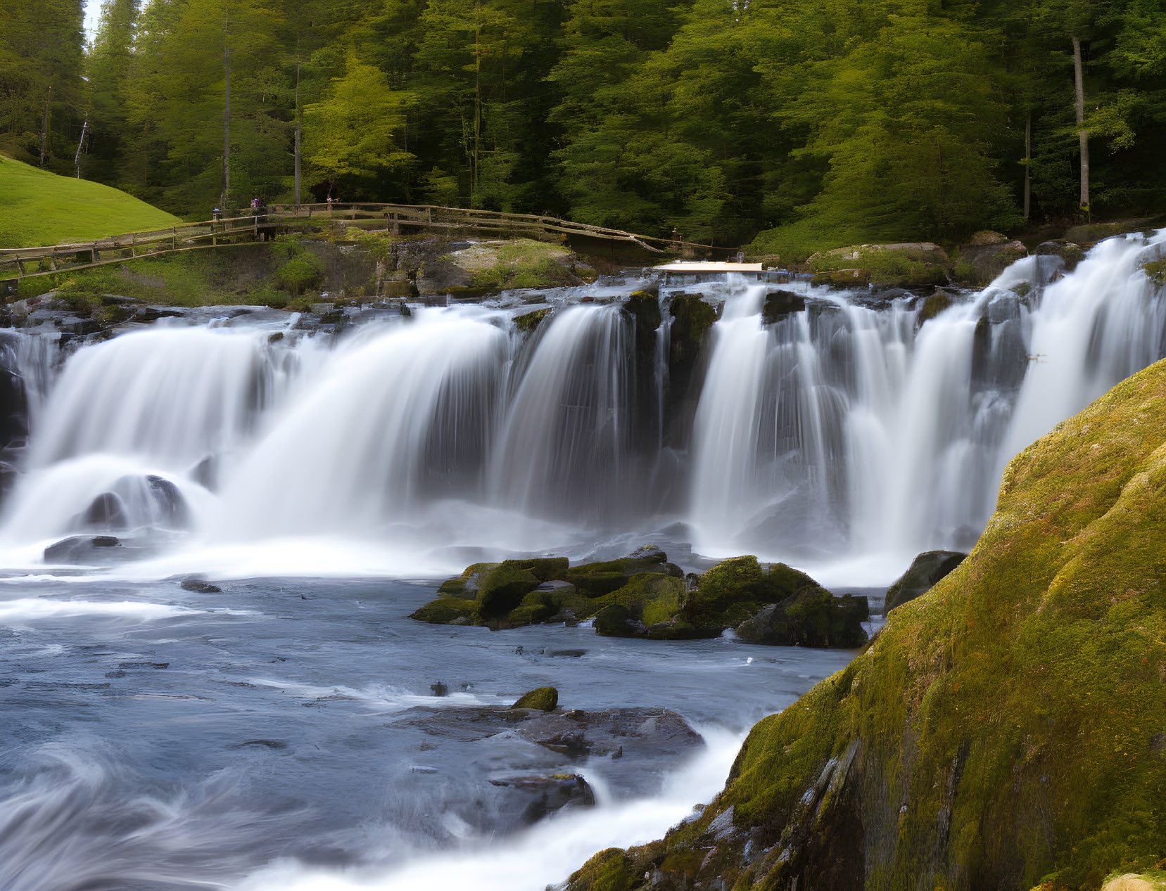 Multiple cascades waterfall surrounded by lush greenery & wooden bridge