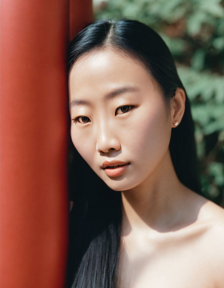 Young woman with dark hair by red pillar, looking at camera in front of green foliage.