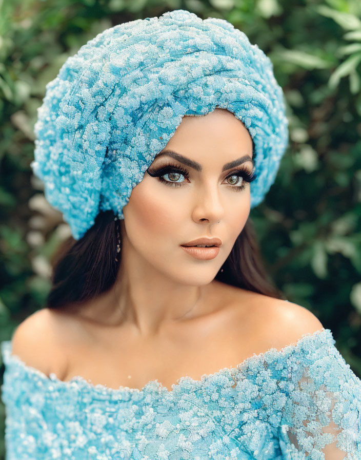 Woman with dramatic makeup in blue lace headwrap and off-shoulder top against green foliage.