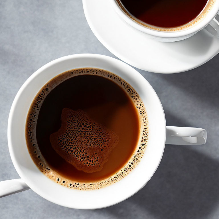 Two frothy coffee cups on grey surface, top view with partial cup.