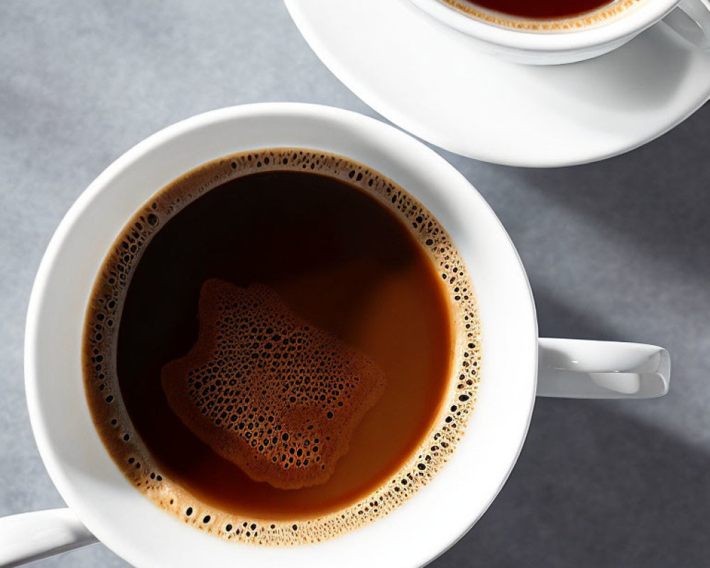 Two frothy coffee cups on grey surface, top view with partial cup.