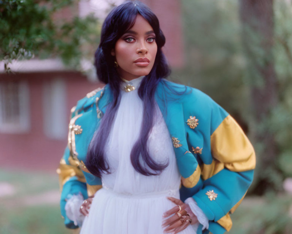 Dark-haired woman in white dress with blue eye makeup and embroidered jacket poses outdoors