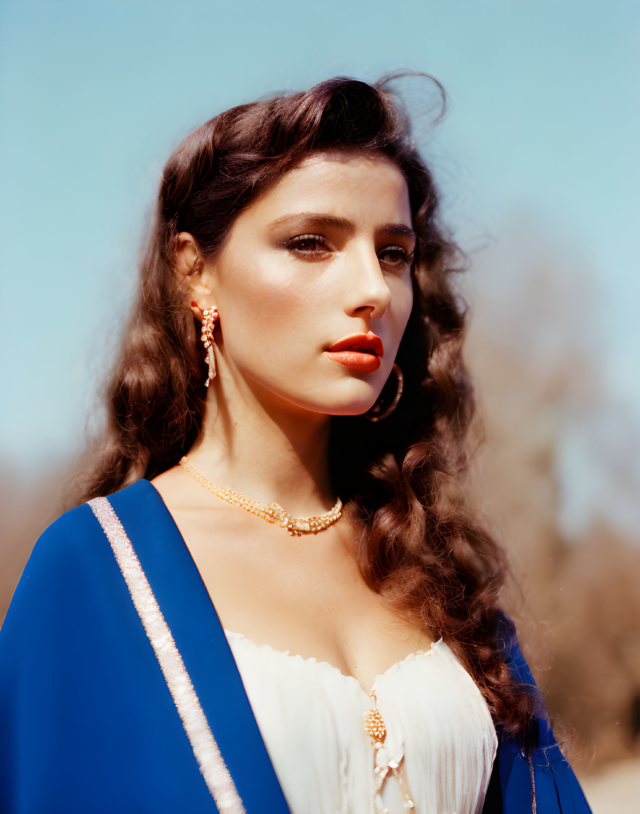 Curly-haired woman in blue and white dress with gold jewelry against blurred background