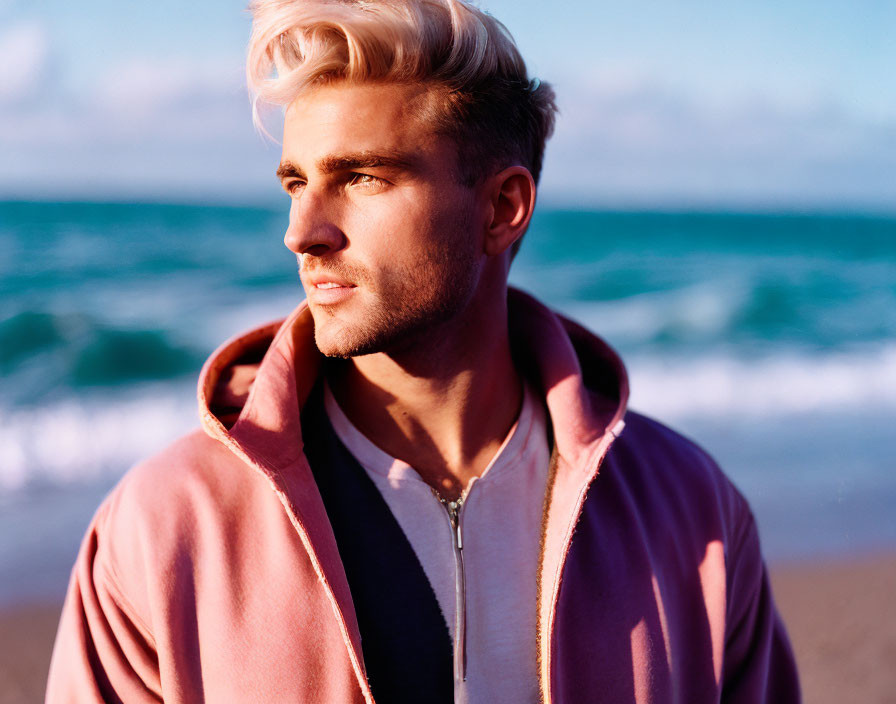 Pinkish-Blonde-Haired Man in Pink and Grey Jacket on Beach with Blue Sky