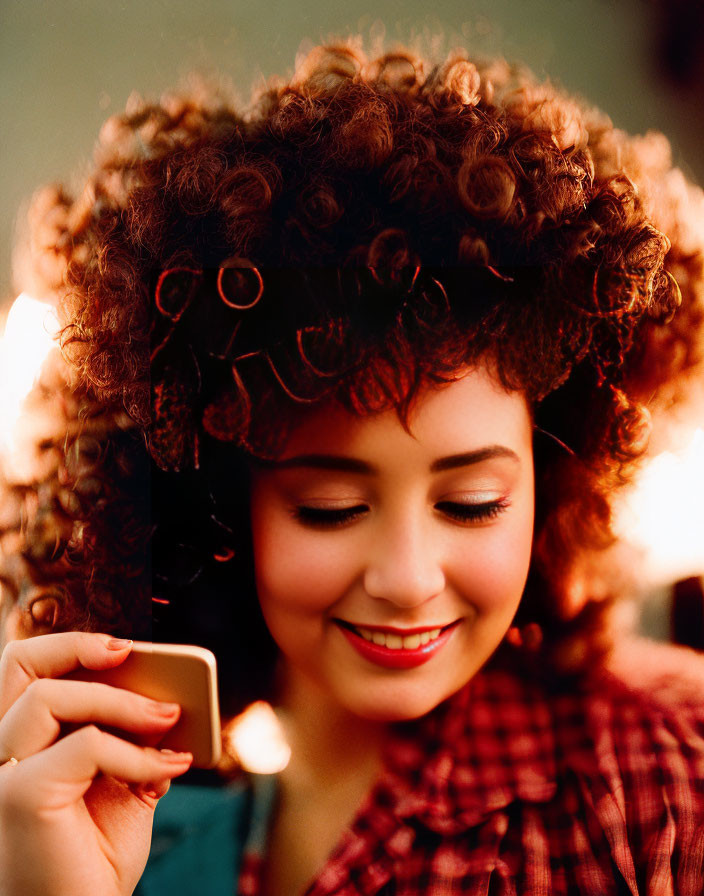 Curly Haired Woman Smiling in Red-Checked Shirt and Looking at Phone