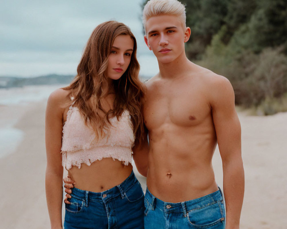 Young man and woman with platinum blonde hair on sandy beach in blue jeans and crop top.