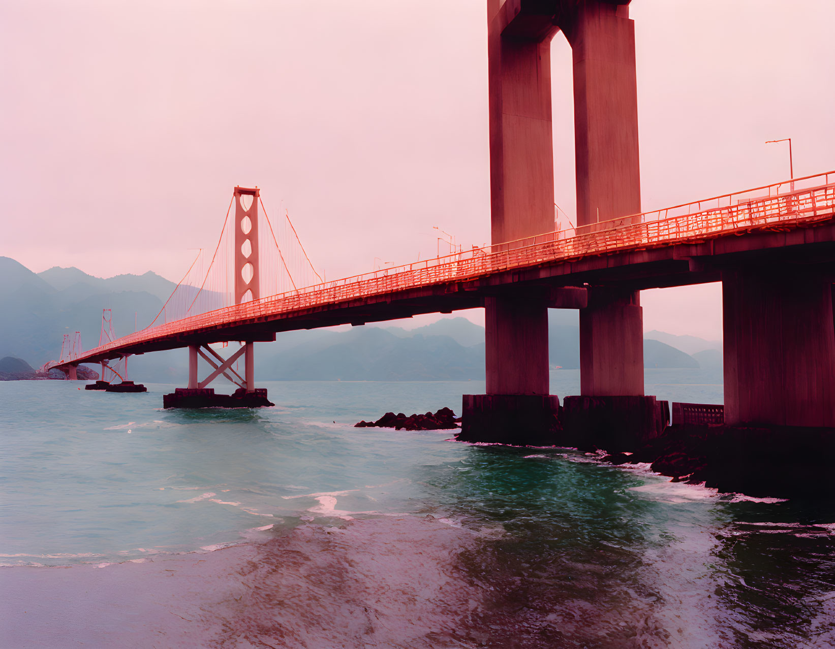 Pink-toned bridge over calm waters with mountains in the backdrop