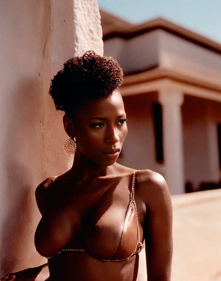 Confident woman with elegant hairstyle in glamorous bikini top and hoop earrings poses by wall