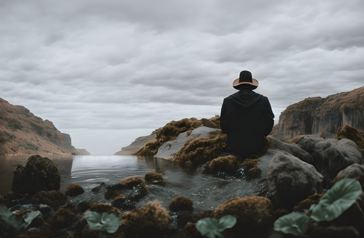 Figure in Black Coat and Hat Sitting by Serene Lake and Mountains