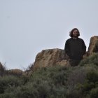 Figure in Black Coat and Hat Sitting by Serene Lake and Mountains