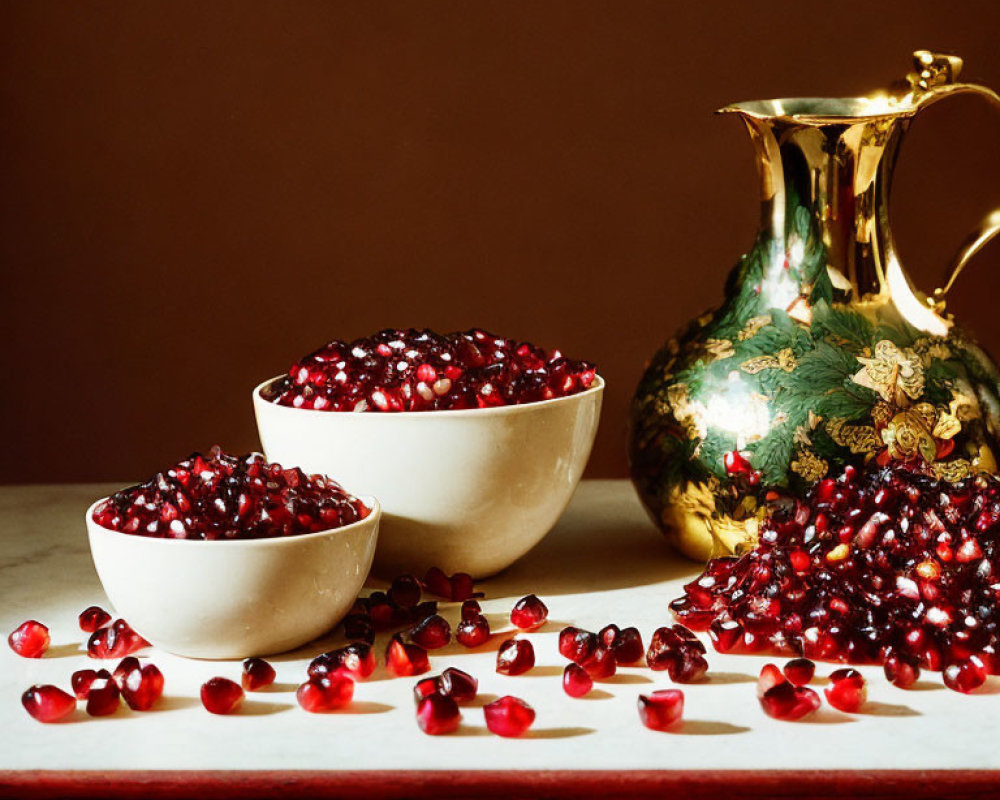 Two white bowls filled with pomegranate seeds beside a gold and green pitcher on a dark table