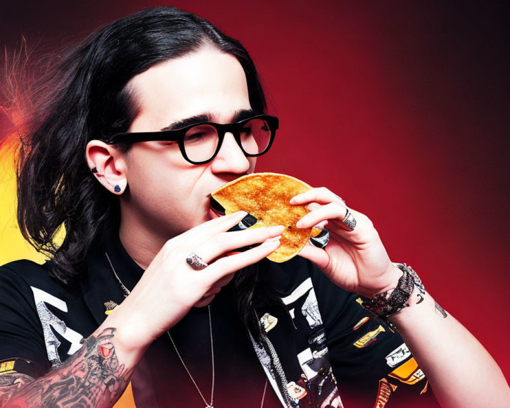 Dark-haired person with glasses and tattoos eating taco on red-yellow background