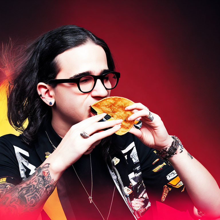 Dark-haired person with glasses and tattoos eating taco on red-yellow background