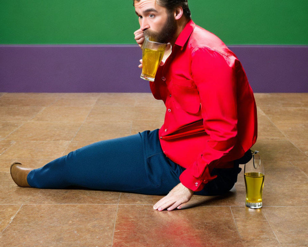 Man in red shirt and blue pants drinking from large glass mug while sitting on floor