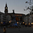 European-style town square at twilight with fountain, buildings, and clock tower.