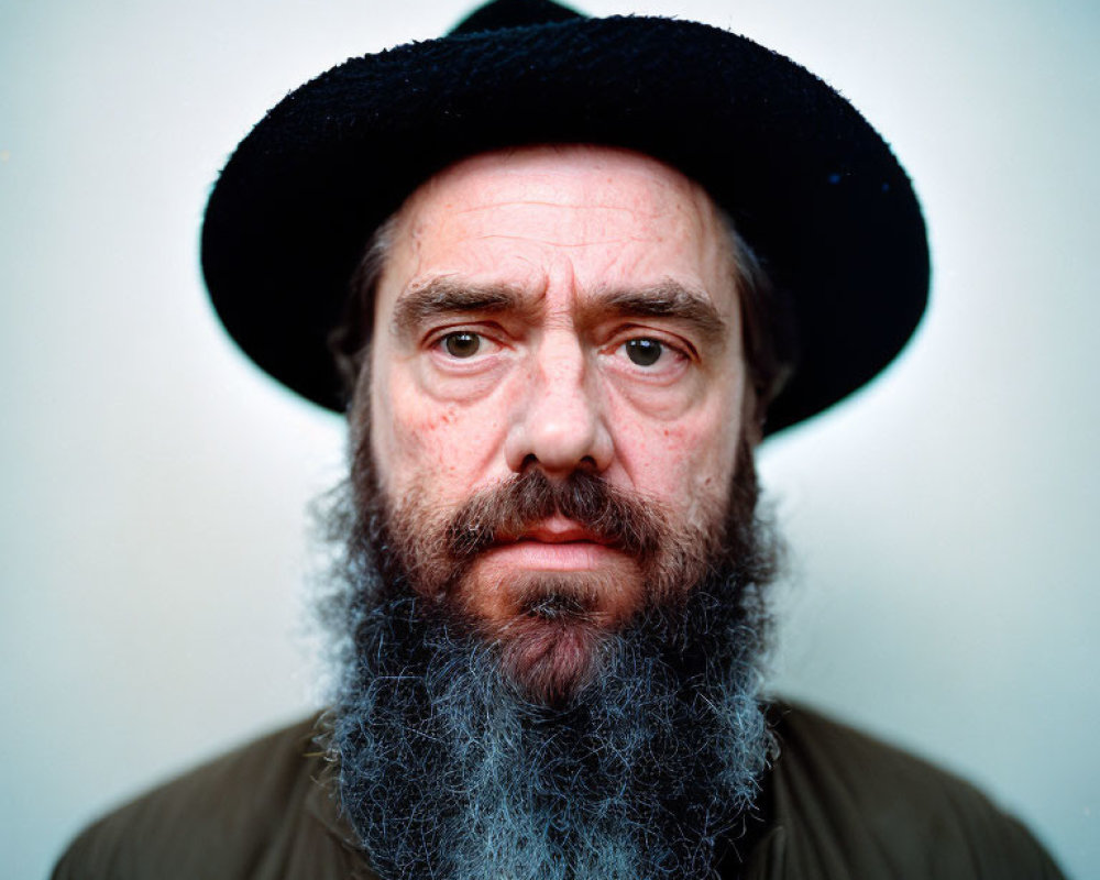 Portrait of man with full gray beard, black hat, and olive green jacket on plain background