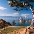 Colorful ribbon-wrapped tree by serene lake with rocky outcrops and mountains under clear sky