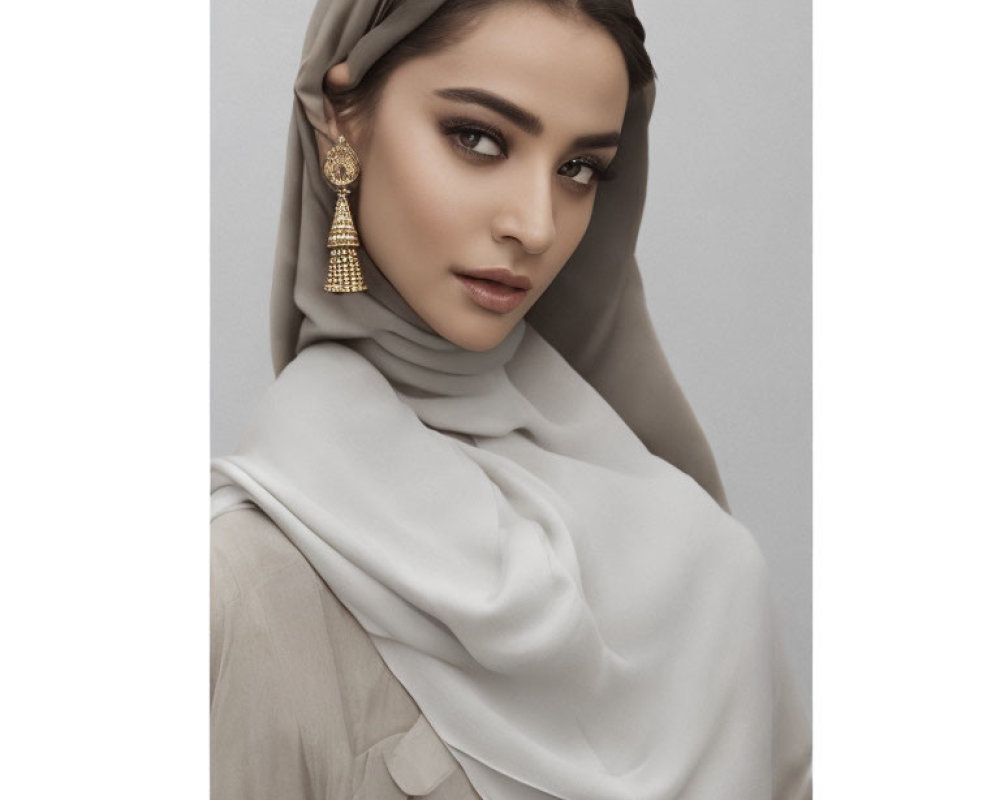 Woman in light-colored hijab and outfit with gold earrings, subtle makeup gazes at camera