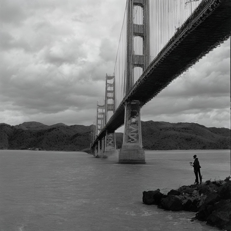 Person standing by water under suspension bridge with pillars, hills, and cloudy sky.