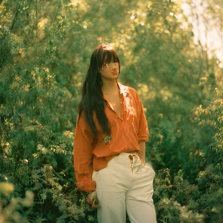 Woman in Orange Blouse and White Pants Surrounded by Greenery