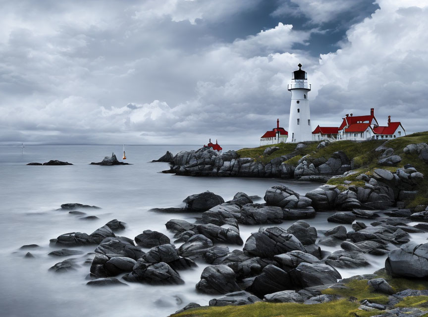 White Lighthouse with Red Rooftops on Rocky Coastal Shore amid Misty Water and Moody Sky