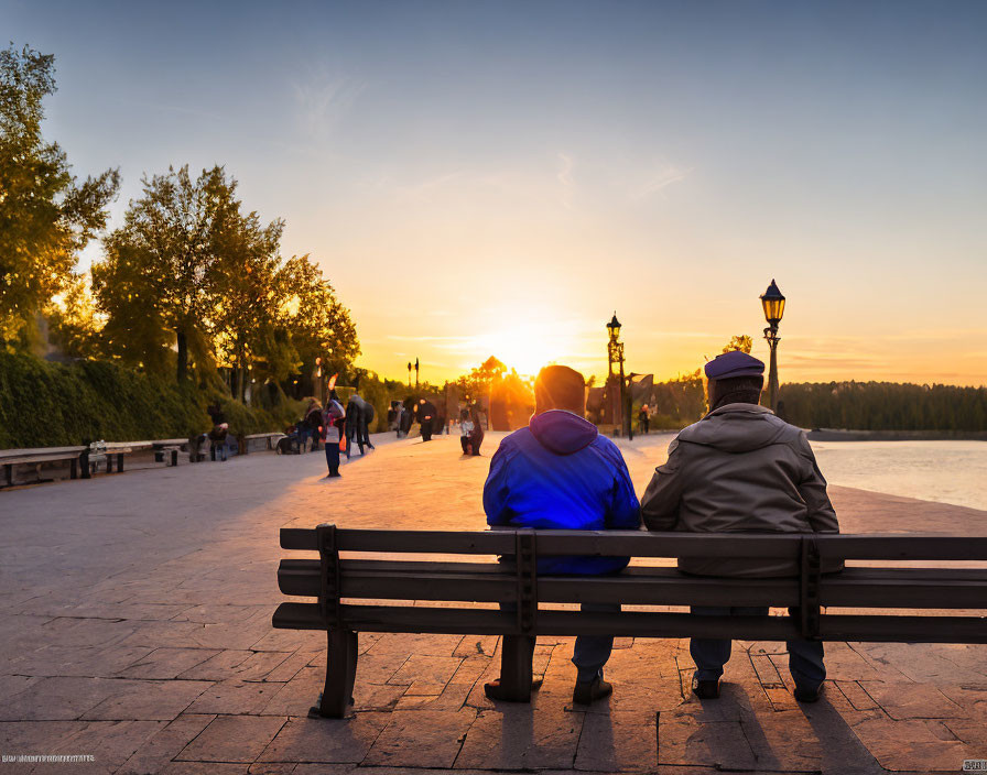 Two individuals on bench watching sunset with trees and path.