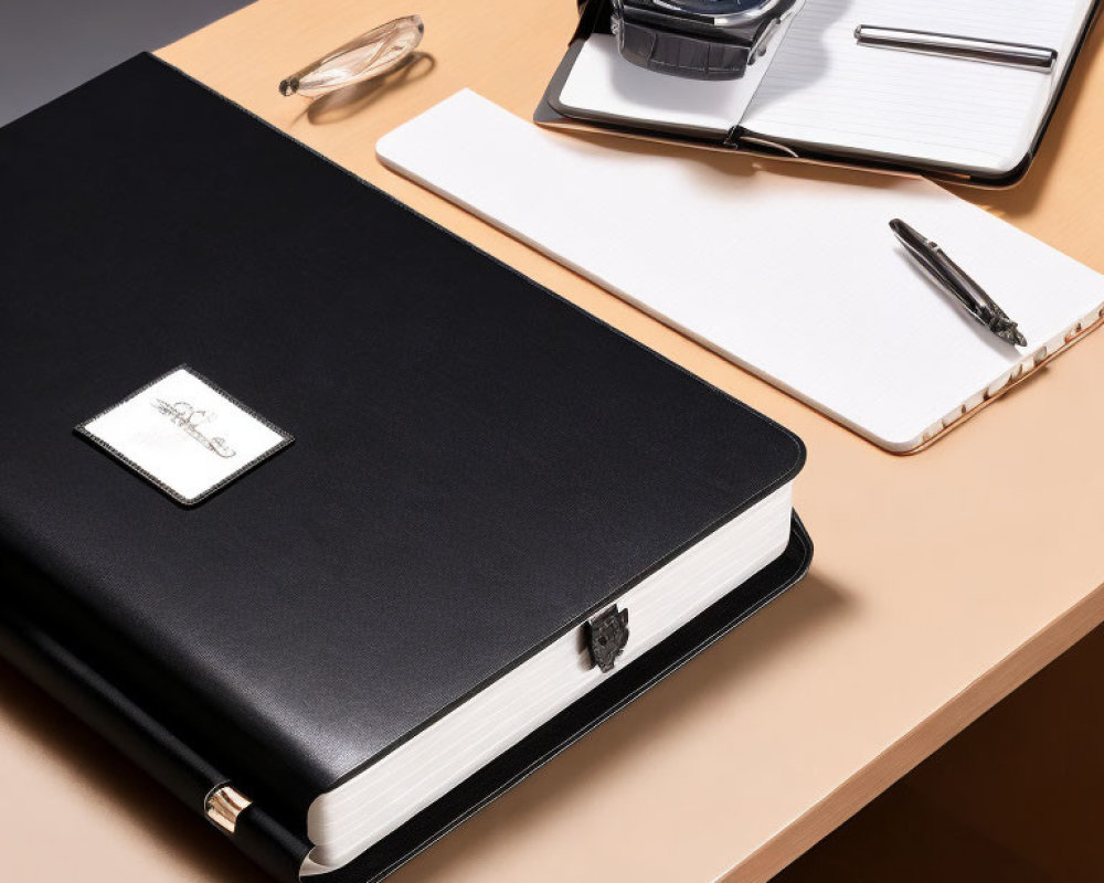 Black Leather Notebook with Silver Emblem, Pen, Wristwatch, and Notepad on Wooden Desk