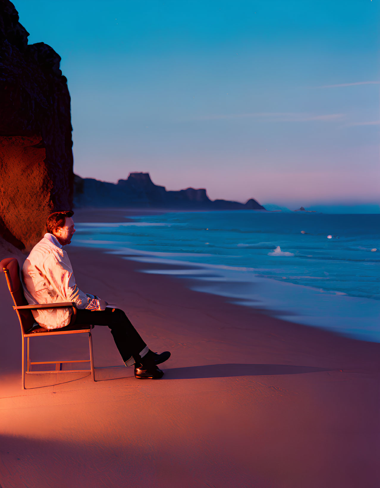 Tranquil beach scene at dusk with person on chair, rocky cliff, and calm ocean waters.