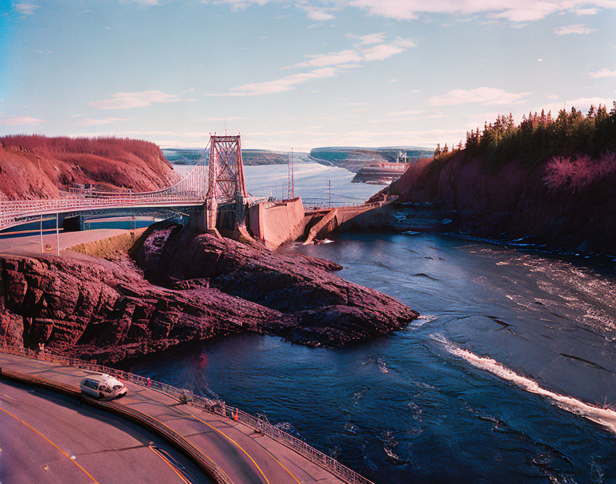 Suspension bridge over rocky river with car on road under pink sky