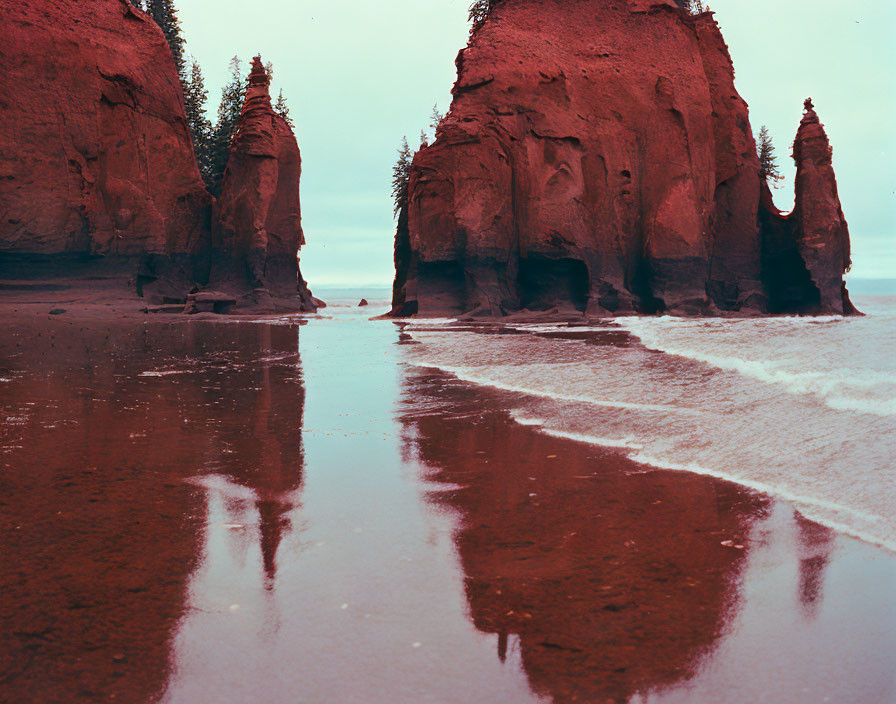 Majestic red sandstone cliffs and rock formations with reflections on wet sand under a cloudy sky.