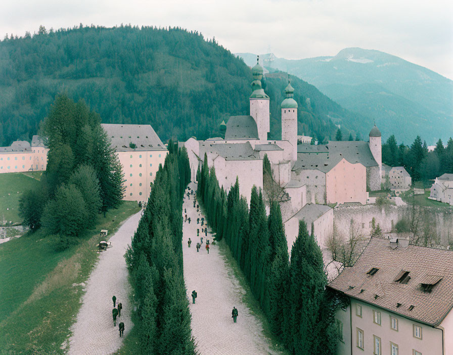 Historic monastery with green domed towers, surrounded by trees, pathway, people, misty mountains