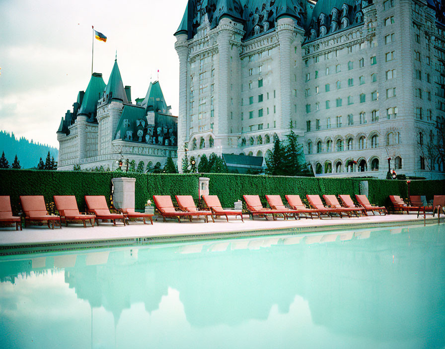 Red lounge chairs by outdoor pool with castle-like building and green hedges under cloudy sky