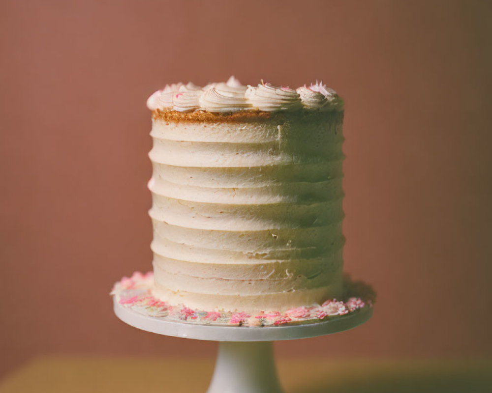 Vanilla cake with white frosting and swirls on white stand, brown background