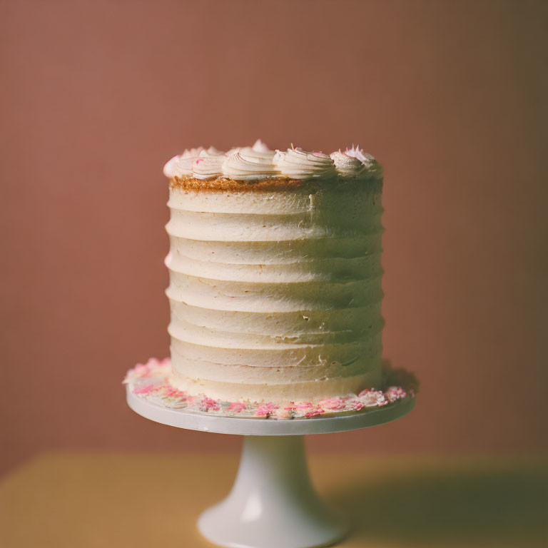 Vanilla cake with white frosting and swirls on white stand, brown background