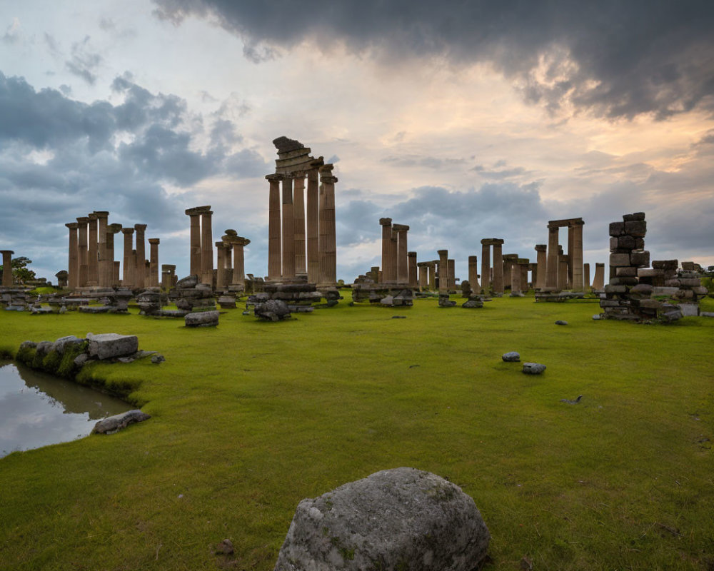 Ancient stone ruins with standing and fallen columns on grassy field under dramatic cloudy sky at dusk