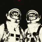 Two astronauts in space suits with helmets, looking to the side, against dark background