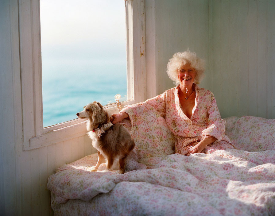 Elderly woman with white curly hair smiles on floral bedspread by ocean view window, petting