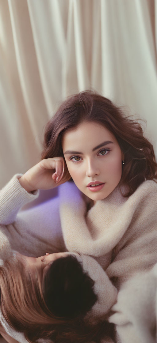 Dark-haired woman in beige sweater leaning on hand against creamy curtain backdrop