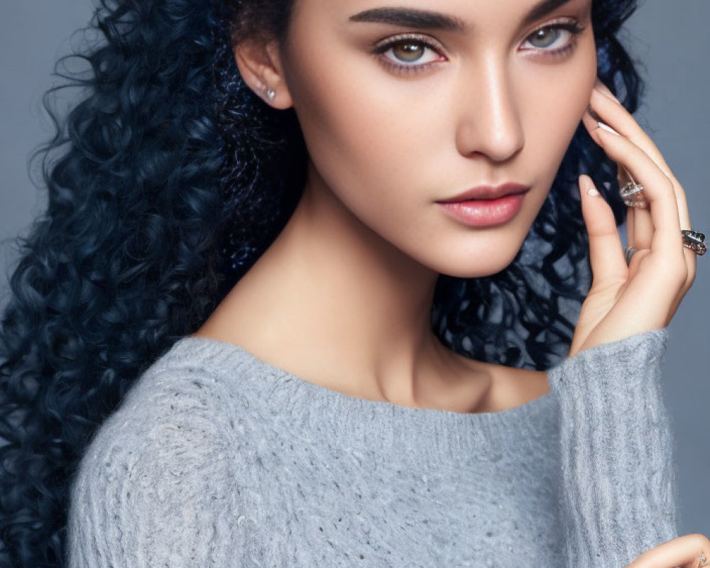 Curly blue-haired woman in grey sweater and rings posing with hand near face